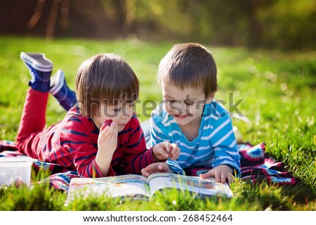 Two adorable cute caucasian boys, lying in the park in a fine sunny afternoon, reading a book and eating strawberries, educating themselves and having fun