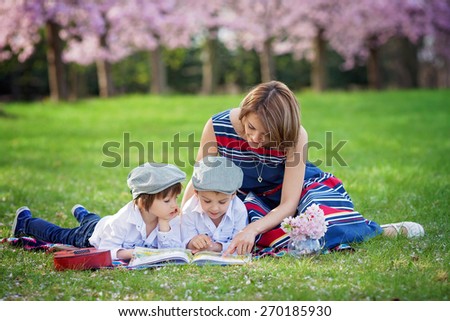 Beautiful portrait of two adorable caucasian boys and their mom, reading a book in a cherry tree blooming garden, spring afternoon, kids lying on the grass on a blanket, vase with flowers and guitar
