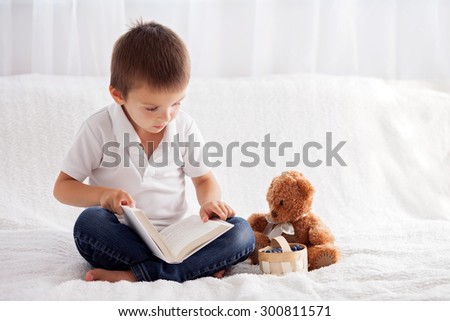 Sweet little boy, reading a book in bed and eating blueberries, teddy bear next to him on the bed