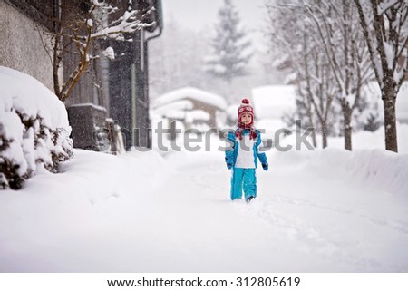 Happy little boy in a snowsuit, hat and scarf, walking through a snowy path with deep snow banks on either side, next to row of houses