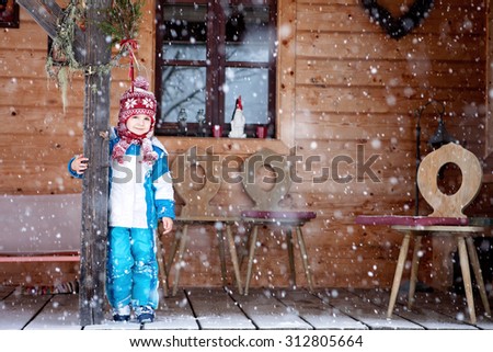 Adorable little boy with lantern, standing in front of a winter cottage while snowing, winter time