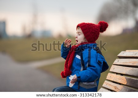 Little boy, sneezing and blowing his nose outdoor on a sunny winter day, sitting on a bench
