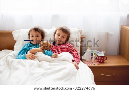 Two sick boys, brothers, lying down in bed with fever, holding teddy bear and resting