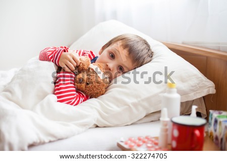 Sick child boy lying in bed with a fever, holding terry bear with band aid, resting