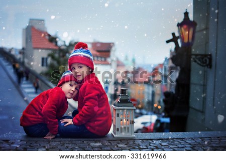 Two cute kids, boys, sitting on stairs, holding a lantern, view of Prague behind them, snowy evening