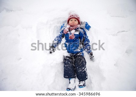 Adorable little boy in blue jacket, red hat and scarf, lying on north pole snow, making snow angel
