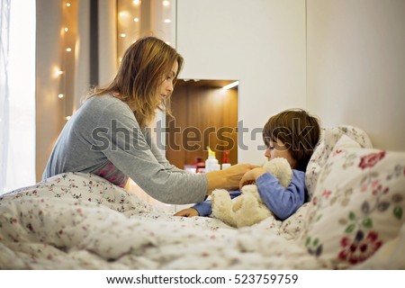 Cute sick child, boy, staying in bed, mom giving him medicine and checking for fever