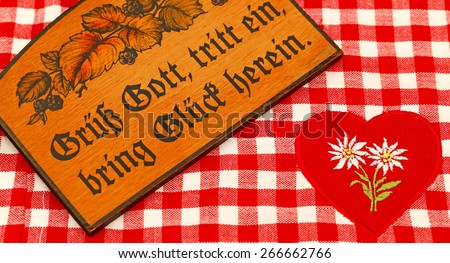 Hello, come in, bring us happiness! in German. Natural  wooden rustic country board with Bavarian greeting on the checkered tablecloth  with a red heart and white embroidered Edelweiss background