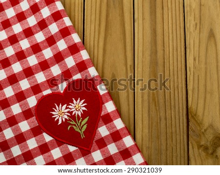 White embroidered Edelweiss flower on heart appliques on the checkered red white tablecloth on wooden table rustic background. Focus on the Edelweiss
