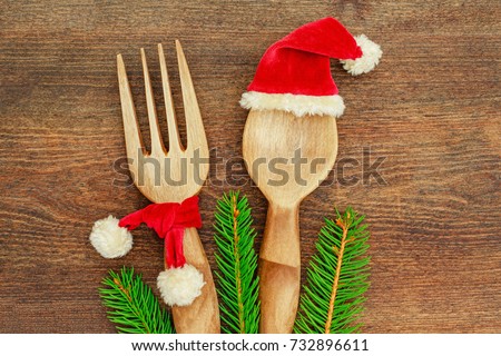 Kitchen wooden spoon in red Santa Claus hat and fork in red Santa Claus scarf on wooden table, top view. Christmas table place setting with cutlery spoon, fork,  green tree branch, decorative clothing