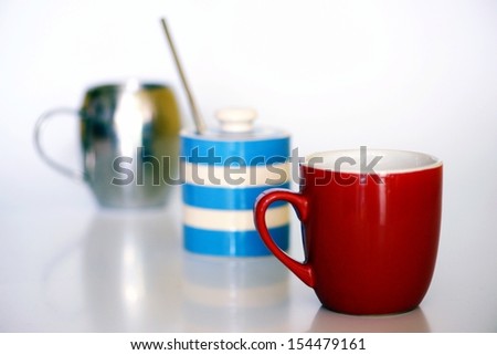 Breakfast coffee mugs in chrome and red with blue and white sugar bowl on white kitchen background with reflections.