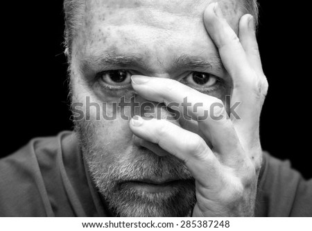 Middle aged man with beard with hand on head and eye looking between fingers as though in migraine pain or anxiety isolated on black background.