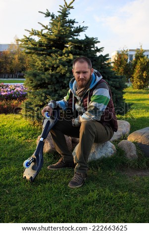 Man relaxing after work-out and sitting outdoors with a skateboard or roller-surf: green fir and grass on the background
