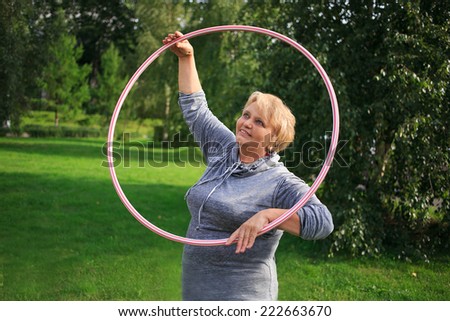 Portrait of happy pretty senior woman exercising with colorful hula hoop on nature background
