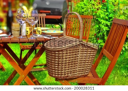 Summer Party or Picnic Scene. Basket, wooden table with glasses and tableware, BBQ Grill in background. Tilt-shift effect.