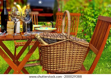Summer Party or Picnic Scene. Basket with sparkling wine, wooden table with glasses and tableware, garden in background with tilt-shift effect.