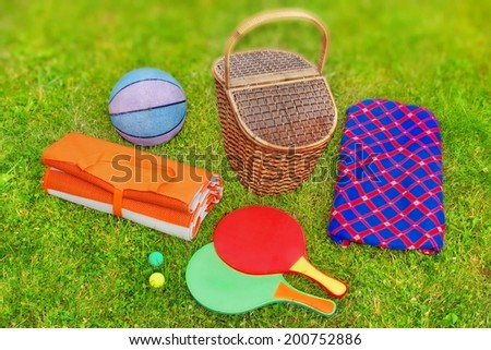 Picnic scene. Basket,  blanket,  racquetball and ball in the grass. Tilt-shift effect in background.