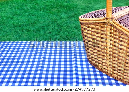 Picnic Basket On The Table With Blue White Checkered Tablecloth. Summer Lawn In The Background