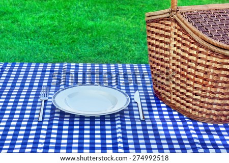 Summer Weekend Picnic Concept. Basket With Food,Empty White Plate,Blue White Picnic Tablecloth, Fork, Knife And Spring Grass In The Background