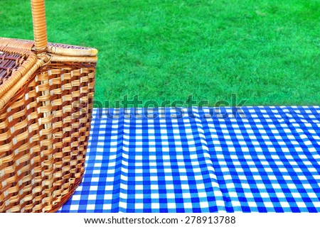 Picnic Basket On The Table With Blue White Checkered Tablecloth. Summer Lawn In The Background