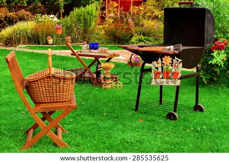 Garden Wooden Furniture, Picnic Hamper Basket, BBQ Grill, Sign Welcome, Wine Glasses On The Table, Plants, Trees and House In The Background. Backyard  BBQ Grill Party Or Picnic Concept