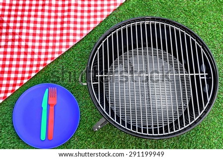 Overhead View Of Red Picnic Tablecloth, Plate, Fork, Knife, BBQ Grill  On The Summer Green Lawn Background