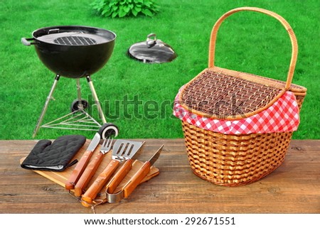 Weekend Summer Outdoor  BBQ Grill Party Ot Picnic Conceptual Scene With Picnic Wood Table, BBQ Tools, Hamper And Open Grill Appliance On The Backyard Lawn In The Background