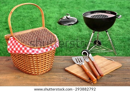 Outdoor Picnic Or BBQ Grill Party Scene At Summertime. Picnic Table With Hamper And Grill Tools Close-up, Barbecue Appliance On The Fresh Lawn In The Background.
