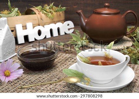 Home Tea Time Scene. Tea Cup With Lime Tree Herbal Green Tea, Honey, Herbs Leafs In The Basket, Clay Teapot, Vintage Spoon And Sign Home On Rustic Old Wooden Table