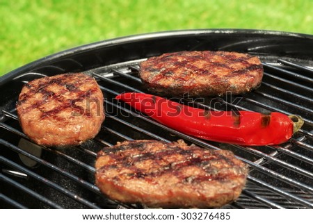 Close-up Of BBQ Hamburger Patties And Chili Pepper On The Hot Charcoal Grill. Vibrant Backyard Lawn In The Background. Cookout Food For Summer Weekend Picnic Or Party.