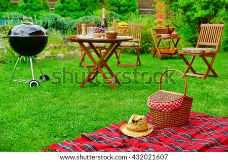 Closeup Of Red Picnic Blanket With Straw Hat And Basket Or Hamper. Blurred Outdoor Wooden Furniture In The Background. Family Home Backyard BBQ Party Or Picnic Conceptual Scene
