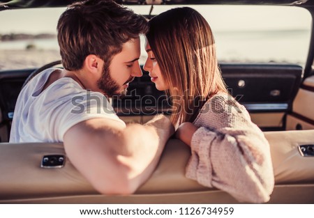 Romantic couple is sitting in green retro car on the beach. Handsome bearded man and attractive young woman in vintage classic car. Stylish love story. Hugging and kissing while being in car.