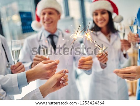 Merry Christmas and Happy New Year! Group of doctors celebrating winter holidays at work. Medical personnel in uniform and Santa Claus hats drinking champagne together.