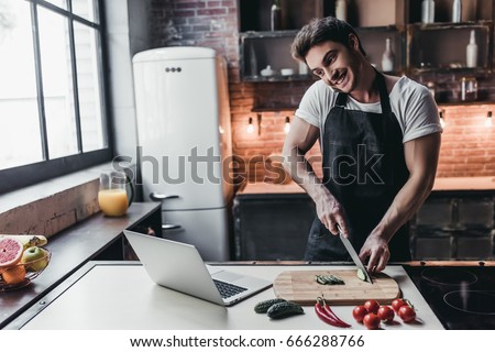 Attractive young man is cooking on kitchen with laptop on table while talking on smart phone.