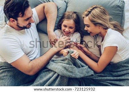 Top view of happy family is having fun in bedroom. Enjoying being together. Parents are tickling their little daughter while lying in bed.
