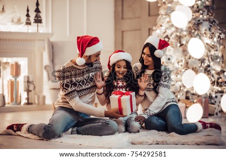 Merry Christmas and Happy New Year! Happy family is waiting for the New Year in Santa Claus hats while sitting near beautiful Christmas tree at home. Parents presenting gift box to charming daughter.