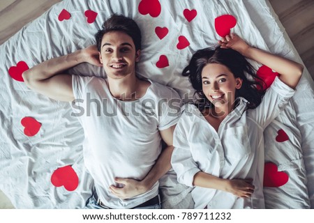 Beautiful young couple in bedroom is lying on bed with red hearts nearby. Enjoying spending time together. Celebrating Saint Valentines Day.