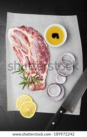 Raw beef steak with rosemary & red onion and chef knife on baking paper & dark wooden background . olive oi l, lemon & balsamic vinegar.