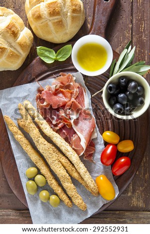 prosciutto crudo with bread, olives, tomatoes, bread sticks, olive oil and basil on old wooden table.