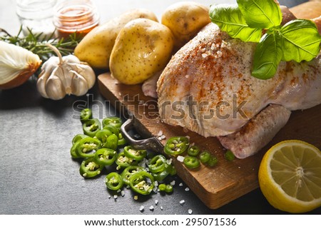 raw whole chicken with garlic potatoes with herbs ready for roasting on a wooden board on a black background