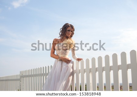 Caucasian model in long white dress posing near white fence outdoors