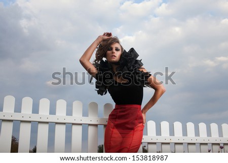 Fashionable model posing on sky background outdoors. Long red and black dress. Red skirt.  black body. volume accessory on his shoulders. black shoes with spikes