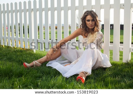 fashionable young woman in long white dress sitting on backyard outdoors. Exclusive beige and white dress. high heels