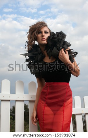 Closeup fashionable model posing on sky background outdoors. hands on waist. Long red and black dress. Red skirt.  black body. volume accessory on his shoulders. black shoes with spikes