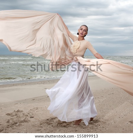 female model in long beige dress posing dynamic in the beach. waving fabric. fluttering long skirt