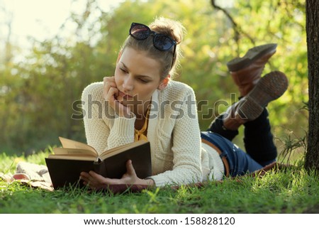 Close Up portrait of reading trendy girl with book lying beneath a tree in the evening