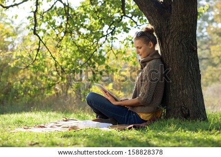 Distance education. Young woman sitting near from tree and working with tablet.