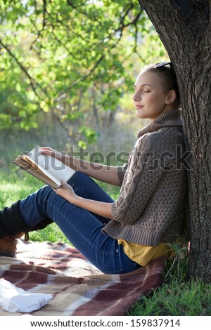 Young woman dreaming with book under the tree during picnic at evening
