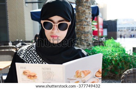 Beautiful arabic woman in a restaurant with the menu in hands. arabic fashion style.\
Middle Eastern woman sitting in a restaurant wearing hijab.