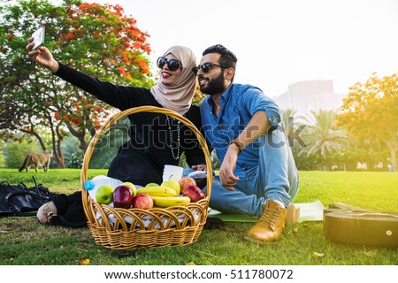 Young beautiful Arabian couple dressed casual and hijab, Abaya, taking a selfie on the lawn in summer park.